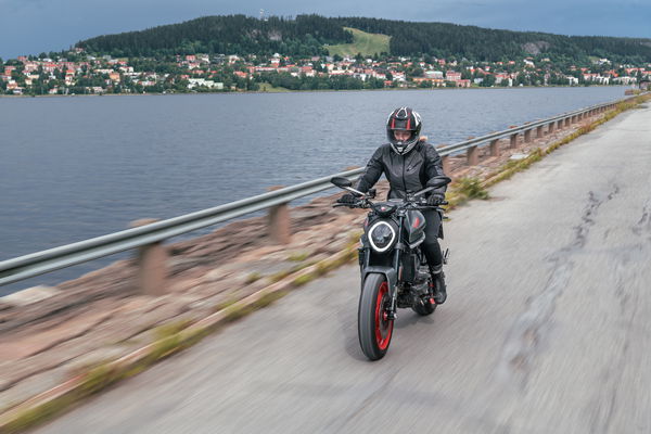 Person with long blonde hair and feminine features on a motorcycle alongside a body of water. Mountains in background.