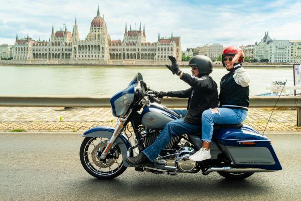 Bill and Cara Davidson riding a Harley-Davidson motorcycle in front of the Hungarian parliament building