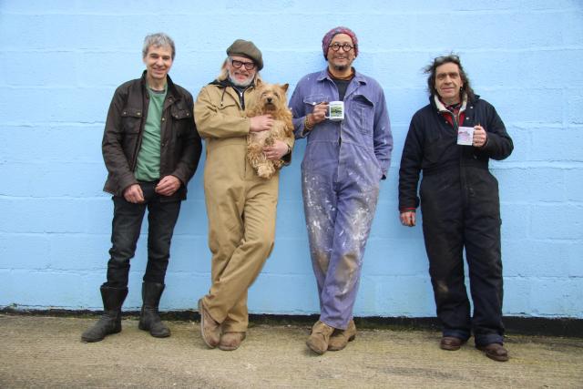 Henry Cole and Friends leaning against a blue brick wall. They're wearing paint statined overalls and are holding mugs of tea and a dog.