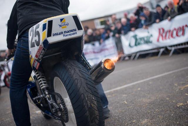 Live Start Up - The rear of a Norton. Flames coming out of the exhaust. There is a man wearing jeans on the bike.