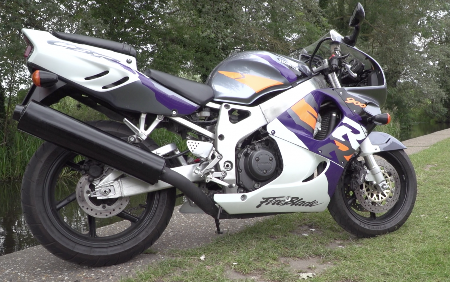 A white, purple, silver and orange 1996 Fireblade CBR900RR parked beside a body of water